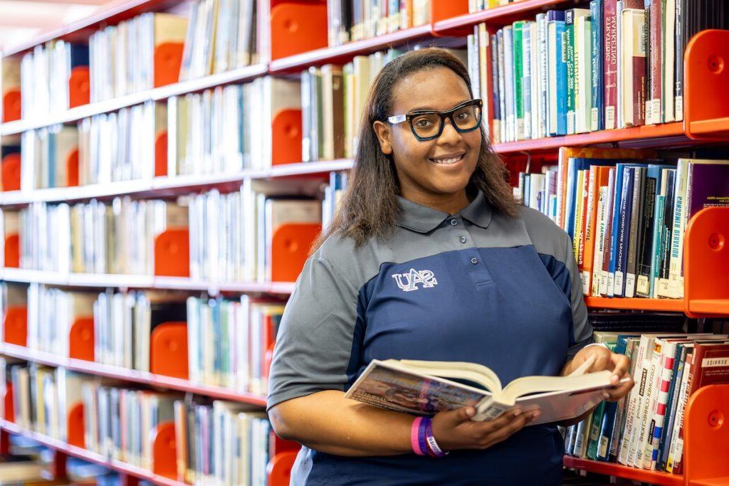 Female student with black rimmed glasses holding an open book, smiling and standing in the library in front of shelves of books